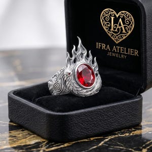 Op de afbeelding: Een zilveren ring met een ovaal geslepen rode edelsteen in een gedetailleerd, vlamvormig metalen ontwerp. De ring wordt gepresenteerd in een zwart sieradendoosje met de gouden tekst IFRA ATELIER JEWELRY en een hartvormig logo op de binnenkant.