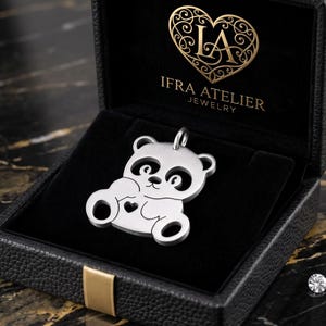 Puede incluir: Un colgante de panda plateado con un pequeño recorte en forma de corazón en el pecho, presentado dentro de un joyero negro texturizado. La tapa interior muestra el texto dorado IFRA ATELIER JEWELRY con un logotipo en forma de corazón.