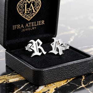 May include: Silver-coloured stud earrings shaped like the letter "R" are presented in an open black jewellery box. The box has a black velvet interior and the brand name "IFRA ATELIER JEWELRY" is printed on the inside of the lid.