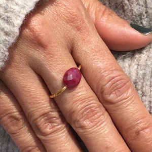 Puede incluir: Un anillo de oro con una piedra roja grande de forma ovalada. El anillo se lleva en el dedo anular de una mano.
