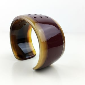 Op de afbeelding: Een bruine en bordeauxrode armband met een gladde, glanzende afwerking. De armband heeft een brede, afgeronde vorm en een licht gebogen ontwerp.