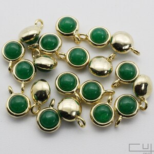 May include: A collection of gold-colored jewelry findings with a round, deep green stone set in the center. Each finding has a small loop for attaching to other jewelry components. The findings are scattered on a white surface.