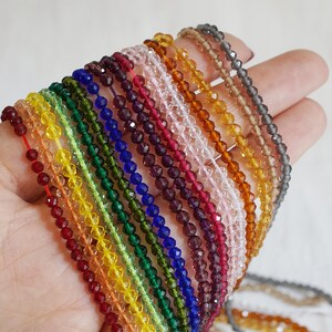 May include: A hand holds multiple strands of small, faceted beads in a spectrum of colours. The beads have a crystalline appearance and are strung on thin cords. Colours include red, orange, yellow, green, blue, purple, pink, and brown. Each strand is a different colour.