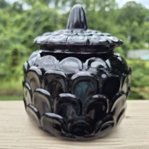 May include: A black glass jar with a matching lid, featuring a raised, repeating scale or petal pattern across the entire exterior surface. The jar has a rounded shape and a pointed finial on top of the lid, with a glossy, reflective finish.