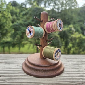 May include: A brown wooden thread spool rack with a circular base and a central post featuring multiple pegs. Three wooden spools of Coats & Clark thread in shades of sage green, dusty pink, and olive green are mounted on the pegs of the rack.
