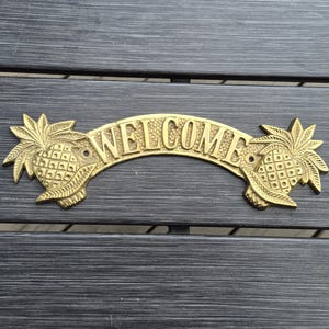 May include: A curved brass welcome sign featuring a pineapple motif on both the left and right ends. The word WELCOME is embossed in capital letters across the center of the arch. The metal has a polished gold-toned finish with visible screw holes.