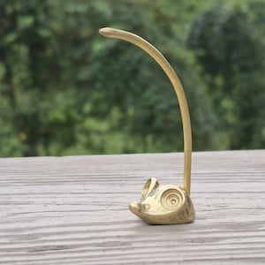 May include: A gold-toned brass figurine shaped like a stylized mouse with a long, curved tail extending upward. The mouse has a smooth, polished surface with circular indentations for eyes and a pointed snout, standing on a flat base.