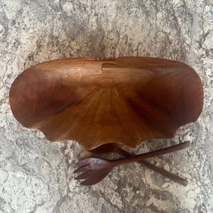 May include: A large, wooden salad bowl with a scalloped edge and two matching salad servers. The bowl and servers are a rich, dark brown color. The bowl is empty and sits on a speckled surface.
