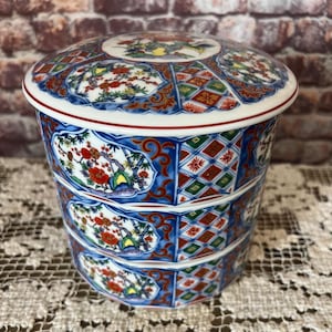 May include: A three-tiered, lidded ceramic container with a detailed floral and geometric pattern. The container features a white rim, red accents, and a predominantly blue, red, and green color scheme. The lid and each tier are decorated with intricate designs.