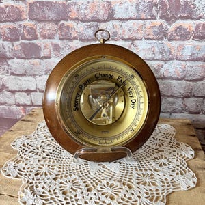 May include: A round analog barometer with a dark wood frame and a gold-toned metal face. The dial features black text reading Stormy, Rain, Change, Fair, and Very Dry, with numerical scales from 28 to 31. A small metal hanging loop is at the top.