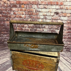 May include: A vintage metal toolbox with a wooden handle, showing signs of rust and wear. The toolbox sits atop a wooden crate with faded red lettering. The background features a brick wall and wooden planks.