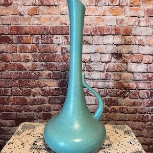 May include: A tall, turquoise-colored ceramic vase with a slender neck and a curved handle. The vase has a bulbous base and a speckled texture. The background features a brick wall. This decorative vase is suitable for floral arrangements or as a standalone art piece.