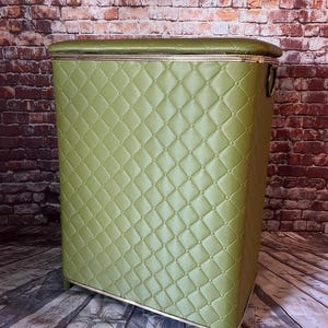 May include: A rectangular olive green laundry hamper featuring a quilted diamond pattern with small circular accents at each intersection. The hamper has a flat lid, gold-toned trim along the edges, and a metal ring handle on the side panel.