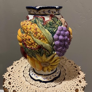 May include: A ceramic vase with a harvest theme, featuring embossed corn, grapes, and sunflowers in vibrant colors. The vase has a white base with blue and gold accents and a decorative rim. It sits on a beige crocheted doily.
