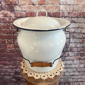 May include: White enamel chamber pot with a lid and a dark blue rim. It has a metal handle and a wooden handle on the side. The pot is sitting on a small wooden stool with a lace doily.