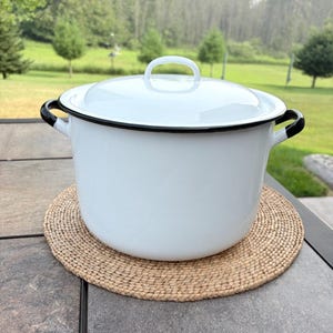 May include: A white enamel stockpot with a matching lid, featuring a smooth glossy finish and a thin black rim along the top edge and the handles. The pot has two side handles and a central handle on the lid, providing a clean look.