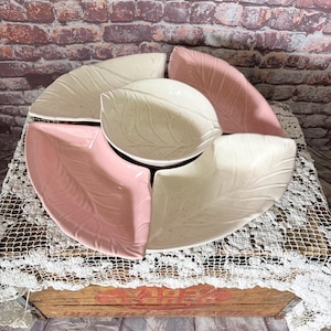 May include: A set of ceramic serving dishes in a leaf design. The set includes a central bowl and four surrounding plates. The plates are in shades of pink and off-white. The dishes are arranged on a white lace tablecloth.