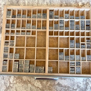 May include: A rectangular wooden letterpress drawer featuring a grid of numerous small compartments. Inside the slots are various metal type blocks, each embossed with individual letters, numbers, and symbols, organized in a structured layout.