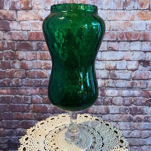 May include: A tall, emerald green glass vase with a bulbous body and a flared rim. The vase sits atop a clear glass stem and a white, crocheted doily. The background features a brick wall.