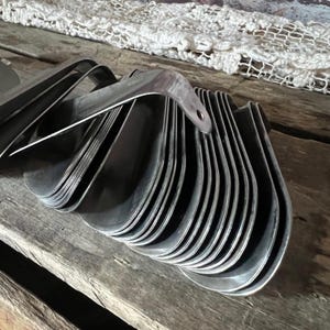 May include: A stack of vintage metal shoe horns, each with a curved handle and a rounded scoop. The shoe horns are made of shiny, silver-colored metal and are arranged on a weathered wooden surface.