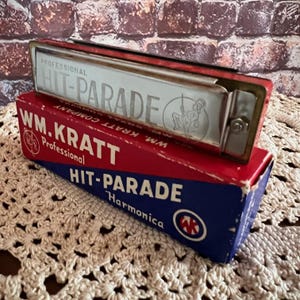 May include: A vintage red and silver harmonica labeled "HIT-PARADE" rests on its original box. The box is red and blue with "WM. KRATT Professional" printed on it. The harmonica has a silver plate with the words "HIT-PARADE" and a graphic of a person playing an instrument.
