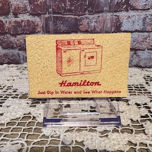 May include: A vintage, rectangular card with a red illustration of a washing machine and dryer set. The word "Hamilton" is printed in red script below the image, with the text "Just Dip In Water and See What Happens" below that.
