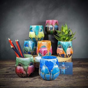 Può includere: Un set di otto vasi in ceramica dipinti a mano in vari colori e design, tra cui blu, verde, rosso e giallo. I vasi sono disposti in gruppo con una piccola pianta grassa in uno dei vasi. I vasi sono decorati con accenti dorati e hanno uno stile unico e artistico.