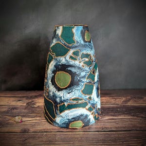 May include: A tall, decorative vase with a unique abstract design. The vase features a pattern of teal, blue, and white shapes outlined in gold. The vase is set against a dark background on a wooden surface.