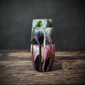 May include: A ceramic vase with a geometric design in shades of pink, black, and green. The vase has a gold trim and a textured surface.