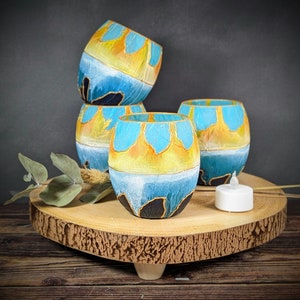May include: Four teal blue and yellow hand-painted candle holders with a wooden base. The candle holders are stacked on top of each other.