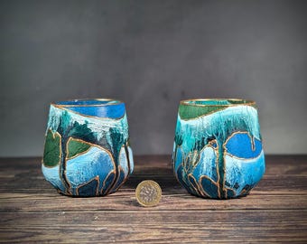 May include: Two blue and green hand-painted ceramic cups with gold accents. The cups are sitting on a wooden surface.