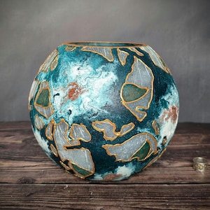 May include: A spherical decorative vase with a teal, blue, and white marbled pattern. Gold-coloured outlines define abstract shapes on the vase. It rests on a wooden surface.