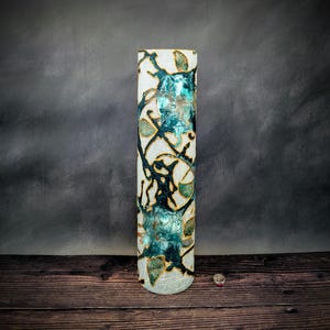 May include: A tall, cylindrical decorative vase with a white background and a teal and gold abstract design. The vase has a crackled texture and is illuminated from within, creating a soft glow. The vase is approximately 30 cm tall.