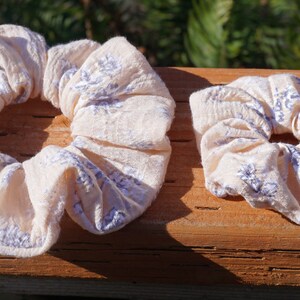 May include: Two fabric scrunchies in a light beige color with a white and purple floral pattern. The scrunchies are on a wooden surface.