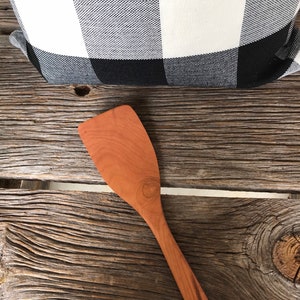 May include: A wooden spatula with a reddish-brown color, lying on a rustic wooden surface. The spatula has a wide, flat blade and a long, slender handle.