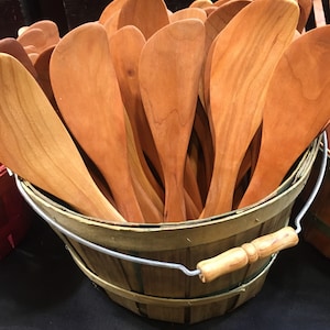 May include: A wooden bucket filled with wooden spoons. The spoons are all made of cherry wood and have a natural finish. The bucket is made of woven wood and has a metal handle.