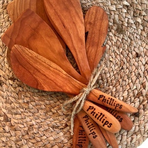 May include: Set of five wooden kitchen spatulas with the name "Phillips" engraved on each. The spatulas are tied together with twine and are sitting on a woven brown surface.