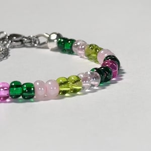 May include: A beaded bracelet featuring a variety of colorful beads. The beads are in shades of green, pink, and purple. The bracelet has a silver clasp.
