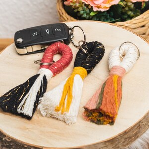 Può includere: Tre portachiavi a nappa fatti a mano in filato nero, bianco e multicolore con accenti dorati. Una chiave dell'auto nera è accanto ai portachiavi su una superficie di legno. Rose color pesca sono in un cestino di vimini sullo sfondo.