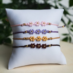Könnte beinhalten: Vier handgefertigte Blumenarmbänder in Rosa, Lila, Beige und Braun, jeweils mit goldenen Akzenten. Die Armbänder werden auf einem weißen Kissen präsentiert, mit grünem Laub im Hintergrund. Die Blumenperlen bestehen aus kleinen, runden Perlen.