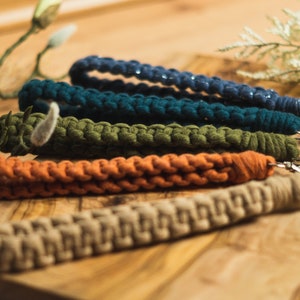 Op de afbeelding: Een verzameling macramé pols keychains in diverse kleuren, waaronder marineblauw, teal, olijfgroen, oranje en beige. Elke keychain heeft een geweven ontwerp en een zilverkleurige sluiting. De keychains liggen op een houten oppervlak.