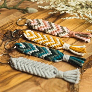 Puede incluir: Cuatro llaveros de macramé hechos a mano en gris, turquesa, amarillo y rosa. Cada llavero presenta un patrón de chevrón tejido y una borla. Se exhiben sobre una superficie de madera con detalles florales.