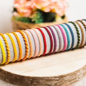 Könnte beinhalten: Eine Sammlung farbenfroher gewebter Armbänder, die auf einer Holzoberfläche präsentiert werden. Die Armbänder sind in verschiedenen Farbtönen erhältlich, darunter Gelb, Orange, Rot, Rosa, Blau und Grün. Einige haben ein geflochtenes Design, andere sind schlicht. Im Hintergrund ein verschwommenes Blumenarrangement.