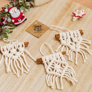 Könnte beinhalten: Drei handgefertigte Makramee-Weihnachtsornamente mit Zimtstangen-Akzenten. Die Ornamente sind cremefarben und haben ein Fransen-Design. Ein Weihnachtsbaum aus Holz und eine Weihnachtsmann-Dekoration sind ebenfalls zu sehen.