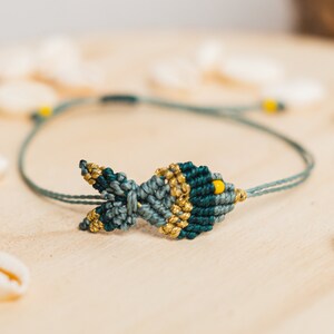 Könnte beinhalten: Ein Makramee-Armband mit Fischmuster. Der Fisch ist aus türkis- und goldfarbenem Garn gefertigt, mit gelben Akzenten. Das Armband befindet sich auf einer hellgrünen Kordel und wird auf einer Holzoberfläche mit Muscheln präsentiert.