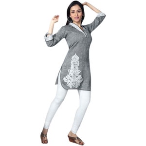 Könnte beinhalten: Eine graue Tunika mit langen Ärmeln und einem weißen, gestickten Blumenmuster. Die Tunika hat einen V-Ausschnitt und einen Kragen. Das Model trägt eine weiße Leggings und braune Schuhe.