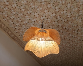May include: A hanging, flower-shaped light fixture made of woven, light-colored material. The fixture is suspended from the ceiling, which has a patterned design in shades of beige and brown. The light is on, illuminating the fixture.
