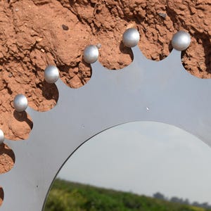 Op de afbeelding: Een zilverkleurige metalen spiegel met een decoratieve kroonachtige rand. De spiegel heeft een reeks afgeronde zilveren studs langs de bovenrand. De achtergrond is een getextureerd, roodbruin oppervlak.
