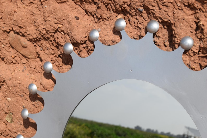 Op de afbeelding: Een zilverkleurige metalen spiegel met een decoratieve kroonachtige rand. De spiegel heeft een reeks afgeronde zilveren studs langs de bovenrand. De achtergrond is een getextureerd, roodbruin oppervlak.