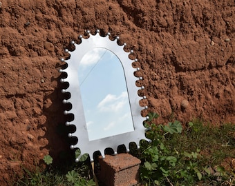 May include: A decorative mirror with a unique, gear-like frame. The silver-colored frame has a series of rounded teeth and small, silver-colored spheres. The mirror reflects a blue sky with white clouds. The mirror is displayed outdoors.
